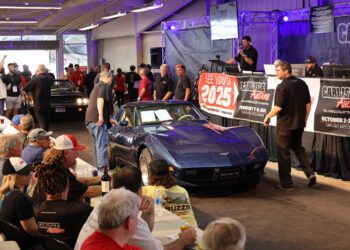 A blue Corvette is presented on stage at the Carlisle Auctions Spring Carlisle Auction, with attendees seated at tables and staff nearby. Signs display auction details and the date for the next Carlisle Auctions event in 2025.