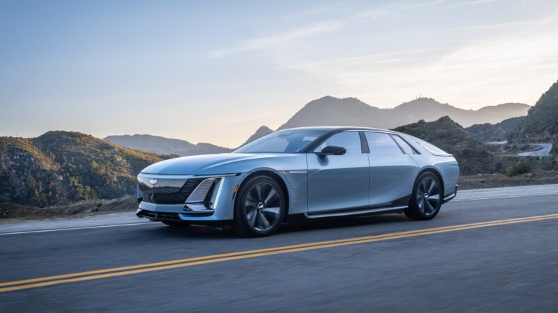 A silver luxury sedan drives on a mountain road with hills and a clear sky in the background.