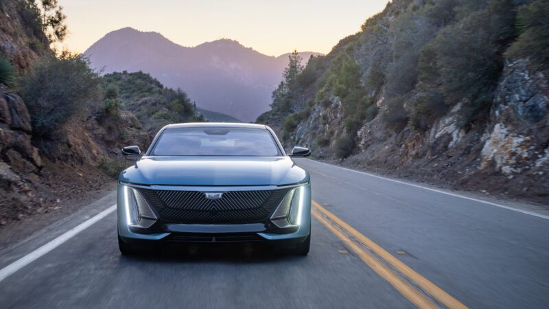 A modern electric car drives on a mountain road at sunset, surrounded by rocky hills and trees under a clear sky.