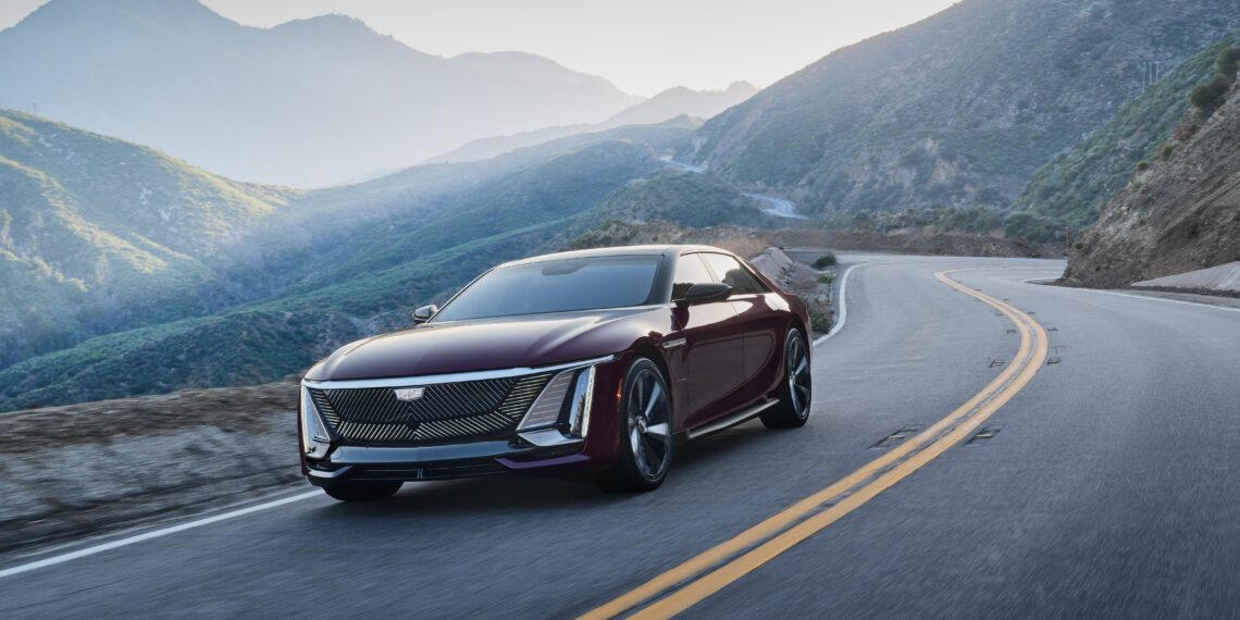 A maroon Cadillac sedan drives on a winding mountain road with green hills and blue sky in the background.