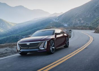 A maroon Cadillac sedan drives on a winding mountain road with green hills and blue sky in the background.