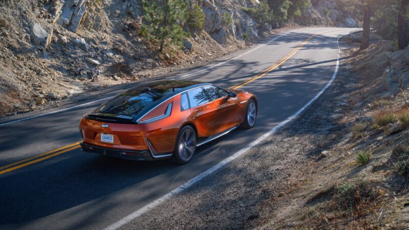 A red Cadillac electric SUV drives on a winding mountain road surrounded by rocks, trees, and sunlight.