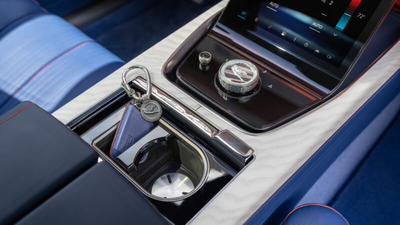 Close-up of a car’s center console featuring a touchscreen, a rotary dial, a key fob with a tag, and a cup holder. Blue and silver interior with red stitching details.