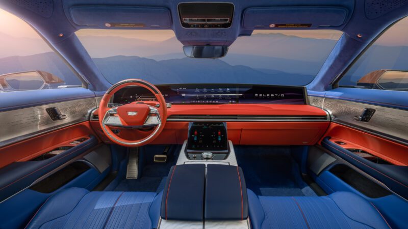 Futuristic car interior with blue and red leather seats, wide digital dashboard display, large steering wheel, and panoramic windshield showing mountains in the background.