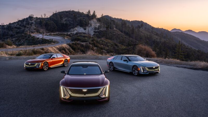Three Cadillac electric cars are parked on a mountain overlook at sunset, with a winding road and forested hills in the background.