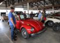 People stand around and observe a red vintage convertible car displayed indoors at the 2025 Spring Carlisle Auction, with other classic vehicles visible in the background at Carlisle Auctions.