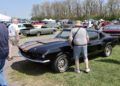 People at a car show observe a black classic Ford Mustang parked on grass, with other vintage cars and tents in the background—reminiscent of highlights from a 2025 Spring Carlisle Auction recap by Carlisle Auctions.