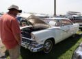 A man in a hat and orange shirt examines the engine of a vintage white car with its hood open at an outdoor car show, capturing the excitement of a 2025 Spring Carlisle Auction event.