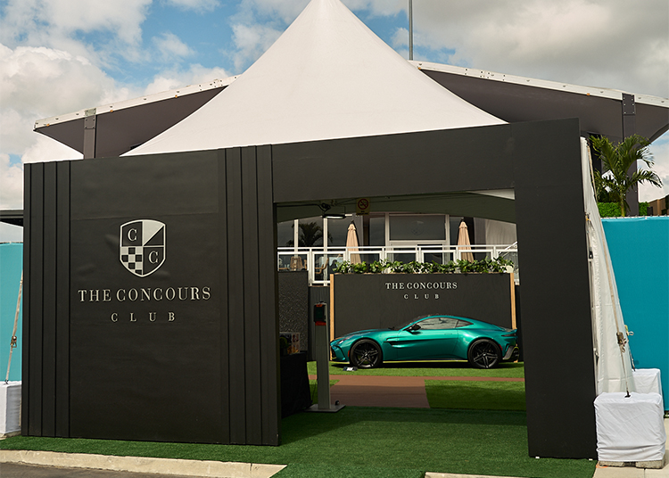 A sleek turquoise sports car is displayed behind a black entrance gate labeled “The Concours Club,” offering a glimpse of the exclusive Luxury Grand Prix Experience, with a white tent and building in the background.