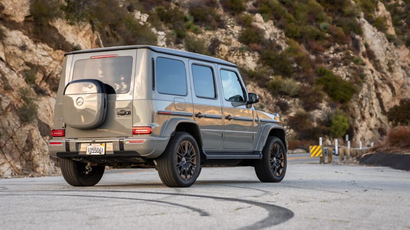 An electrified metallic gray Mercedes-Benz G-Class SUV is parked on a winding mountain road, surrounded by cliffs and lush vegetation.