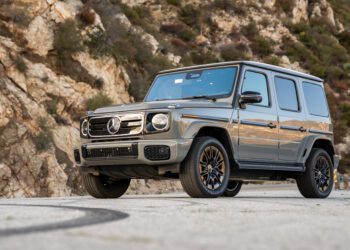 A silver Mercedes-Benz G-Class SUV, a true Mercedes icon, is parked on a paved road with rocky hills and greenery in the background.
