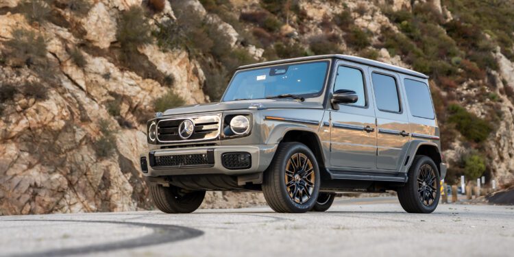 A silver Mercedes-Benz G-Class SUV, a true Mercedes icon, is parked on a paved road with rocky hills and greenery in the background.