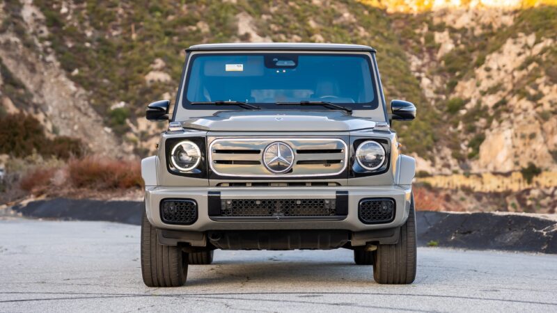 A front view of a gray, electrified Mercedes-Benz G-Class SUV parked on a paved road with hills and vegetation in the background.