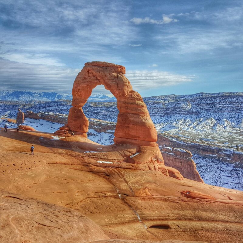 Delicate Arch in Arches National Park, Utah, stands on red rock with snow-dusted mountains behind—a stunning view in one of the top U.S. off-roading destinations—under a partly cloudy sky.