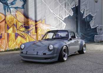 A gray classic sports car from Classic 9 Motorwerks is parked on a street in front of a building with colorful graffiti and a monochrome mural, evoking the vibrant energy of an exotic car festival.