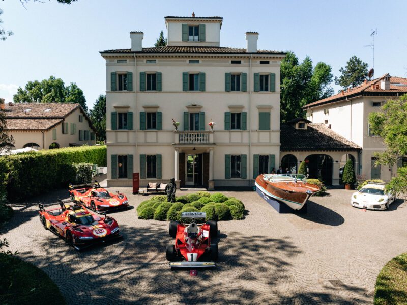 Five vintage sports cars and a classic wooden speedboat are displayed in front of a grand villa with green shutters, celebrating Best of Show Winners at the Cavallino Classic Modena 2025 on a sunny day.
