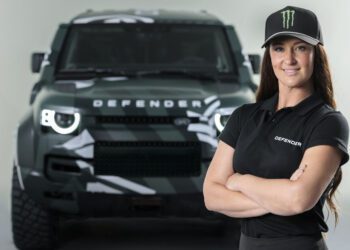 Sara Price stands confidently with arms crossed in front of a green Land Rover Defender, ready to take on any challenge like the Dakar Rally.