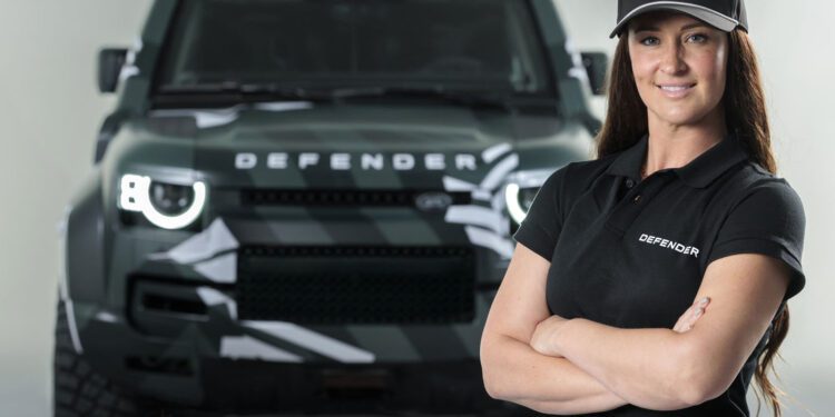Sara Price stands confidently with arms crossed in front of a green Land Rover Defender, ready to take on any challenge like the Dakar Rally.