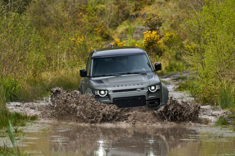 A dark green Land Rover Defender drives through a muddy water-filled path surrounded by greenery, showcasing expedition elegance as it splashes mud and water around its front wheels.