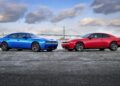 A blue and a red Four-Door Dodge Charger Daytona are parked side by side on an outdoor lot, with a bridge and cloudy sky in the background—proving there’s more to this muscle car sedan than meets the eye.