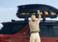 A woman closes a rooftop cargo box on top of a red SUV, with luggage visible inside, echoing the adventurous spirit of Land Rover Marks 35 Years Of Discovery With Gemini And Tempest Special Editions, all on a clear day.