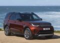 A red Land Rover Discovery SUV, celebrating Land Rover Marks 35 Years Of Discovery With Gemini And Tempest Special Editions, is parked on a dirt overlook near the ocean, with waves and cliffs in the background under a clear sky.