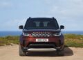 A front view of a maroon Land Rover Discovery, celebrating 35 years with the Gemini and Tempest Special Editions, parked on a sandy area with vegetation and the ocean in the background.