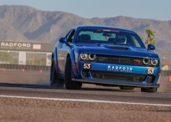 A blue Dodge sports car, number 53, races on a track at Radford Racing School, with mountains and the school's entrance sign visible in the background.