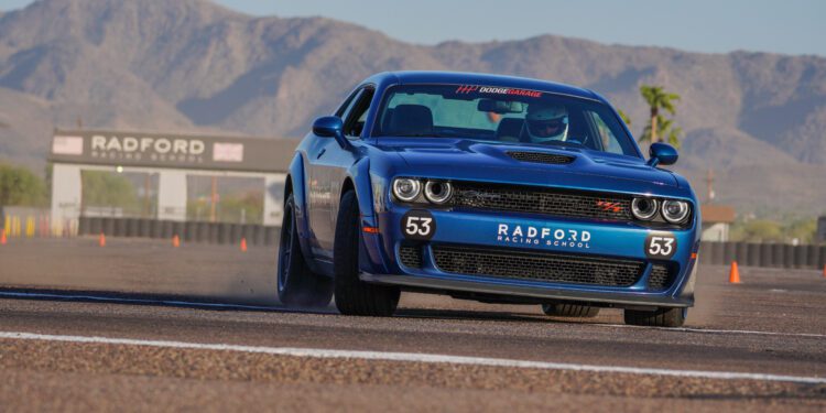 A blue Dodge sports car, number 53, races on a track at Radford Racing School, with mountains and the school's entrance sign visible in the background.