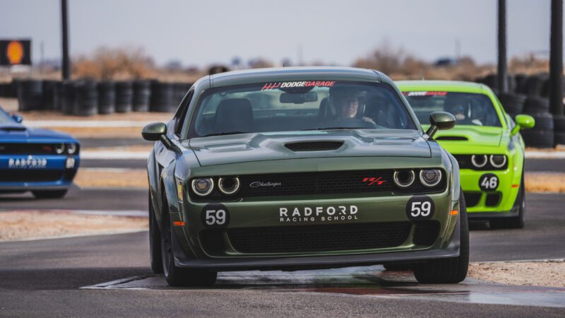 A green Dodge Challenger with the number 59 and "Radford Racing School" on the front is driving on a racetrack, followed by a bright green car and a blue car.