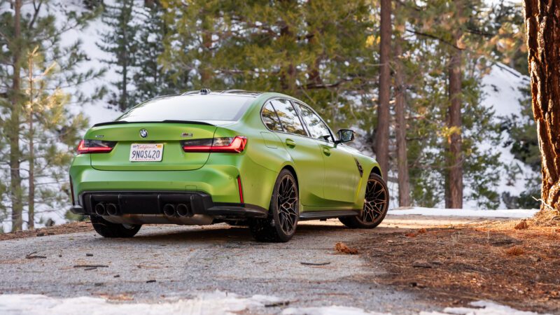 A green BMW sedan with black wheels is parked on a snowy, forested road with pine trees in the background.