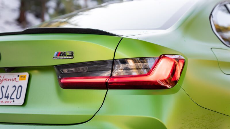 Close-up of the rear left side of a green BMW M3 Competition, showing the taillight, badge, and part of a California license plate.