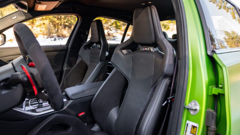 Interior view of a green car showing black leather sports seats with "M3" logo, a steering wheel with red accents, and the passenger door open.