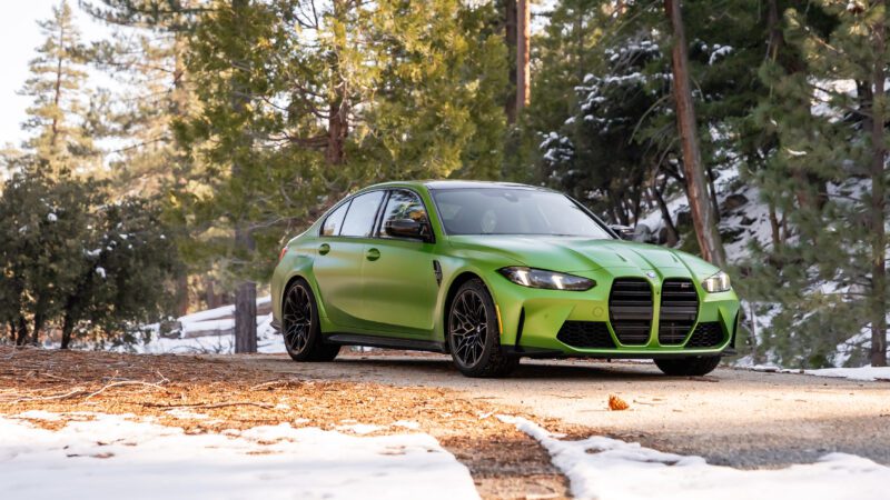 A green BMW sports sedan is parked on a paved road surrounded by trees and patches of snow.