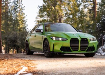 A green BMW sedan is parked on a forested road with trees and patches of snow in the background.