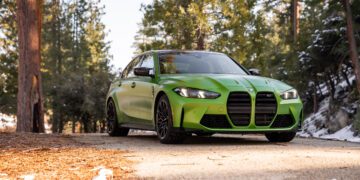 A green BMW sedan is parked on a forested road with trees and patches of snow in the background.