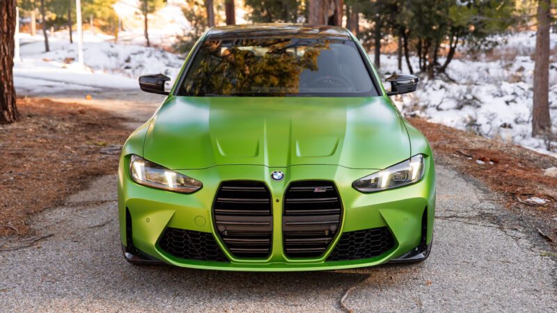 A green BMW sports car is parked on a forest road, viewed from the front, with trees and patches of snow in the background.