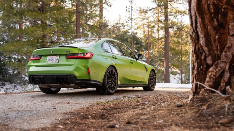 A green BMW sedan is parked on a forest road with trees and patches of snow in the background.