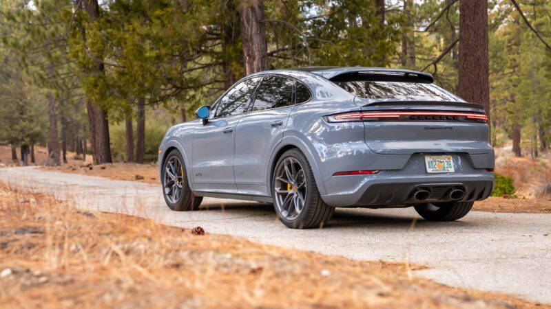 A gray Porsche Cayenne coupe is parked on a narrow paved road in a forested area with pine trees and dry ground.