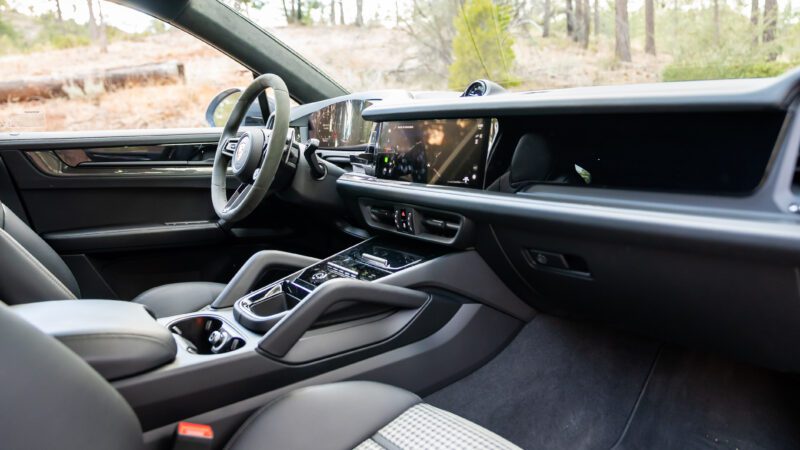 Modern car interior featuring a digital display, touchscreen controls, leather seats, and a steering wheel, with trees visible through the windows.