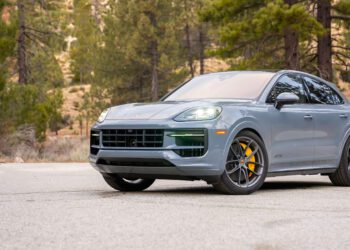 A gray Porsche SUV with yellow brake calipers is parked on a paved surface, surrounded by trees and a forested background.