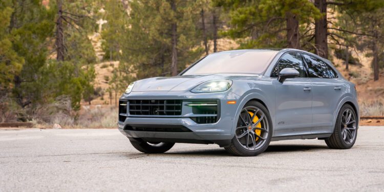 A gray Porsche SUV with yellow brake calipers is parked on a paved surface, surrounded by trees and a forested background.