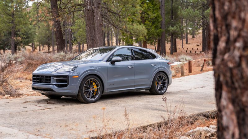 A gray Porsche Cayenne with yellow brake calipers is parked on a concrete path surrounded by trees in a forest setting.