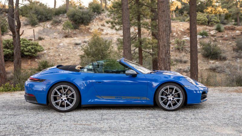 A blue convertible sports car parked on a gravel road in a wooded area with trees and shrubs in the background.