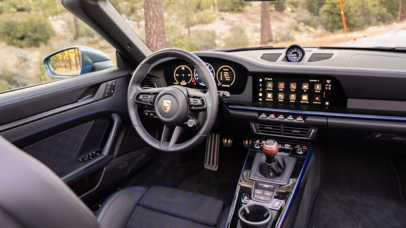 Interior view of a Porsche sports car featuring a leather-wrapped steering wheel, digital displays, touchscreen controls, and a manual gear shift.