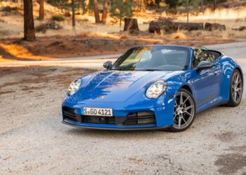 A blue convertible sports car is parked on a gravel road in a wooded, dry landscape with scattered trees and sunlight.