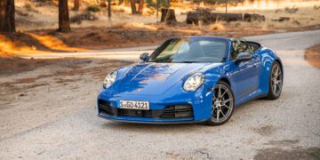 A blue convertible sports car is parked on a gravel road in a wooded, dry landscape with scattered trees and sunlight.