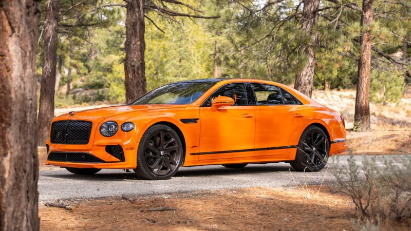An orange Bentley sedan with black wheels is parked on a narrow road in a forested area with tall pine trees.