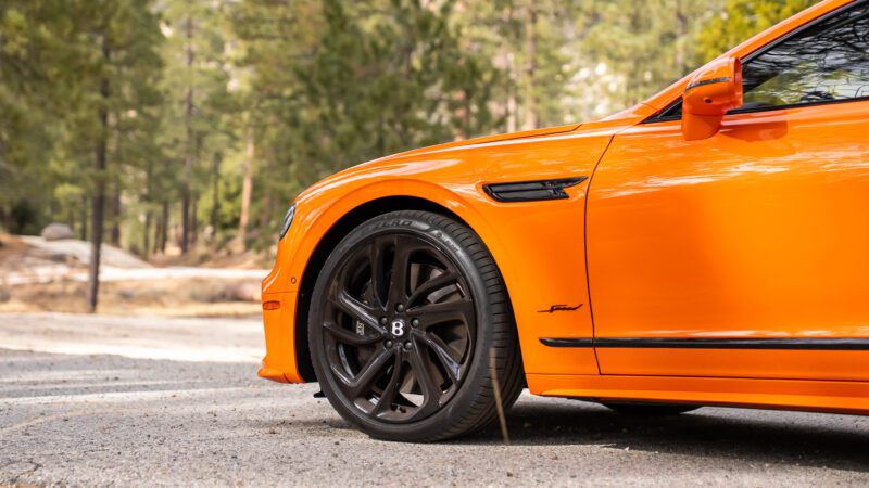 Front side view of an orange luxury car parked on a road, showing the front wheel, side mirror, and part of the hood, with a forest background.