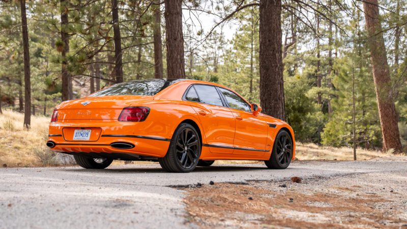 An orange luxury sedan with black wheels is parked on a forest road surrounded by tall trees.